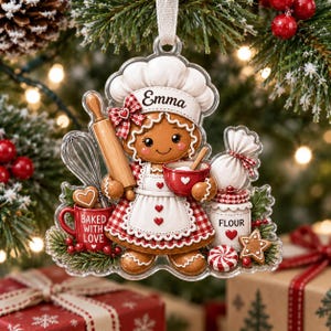 May include: A gingerbread person ornament wearing a chef hat with the name Emma, a red and white checkered apron, and holding a rolling pin and a red bowl. The ornament features a red mug labeled Baked With Love, a flour jar, and a whisk.