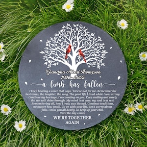 May include: A round, dark gray memorial stone with white text and a tree design. Two red cardinals perch on the tree branches. The text reads "Grandma Albert Thompson 1940-2025" and a poem. The stone is set in green grass.
