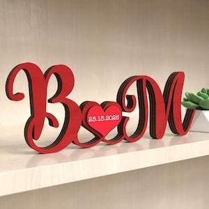 May include: A red wooden decorative piece featuring the letters "B" and "M" with a heart in between. The heart has the date "25.15.2025" printed on it. The letters are in a cursive style and sit on a shelf.