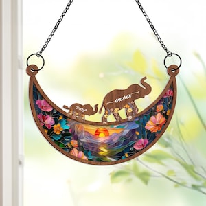 May include: A wooden crescent moon-shaped wall hanging with a stained glass design of a sunset landscape, flowers, and two elephants. The larger elephant has the word "mama" written on it, and the smaller elephant has the word "Freya" written on it.