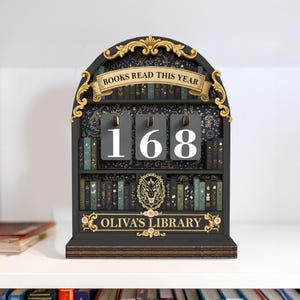 May include: A black wooden bookshelf-shaped counter with the text "BOOKS READ THIS YEAR" and the number "168" displayed on gray flip cards. The bottom of the counter reads "OLIVA'S LIBRARY". The bookshelf is decorated with gold accents and small pink flowers.