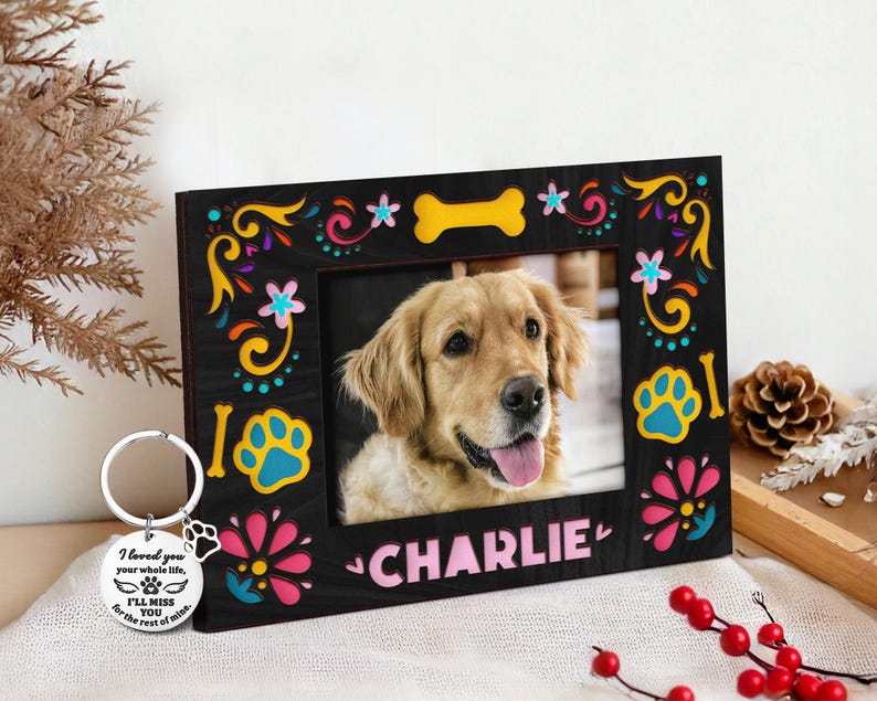 May include: A black wooden picture frame with a photo of a golden retriever dog named Charlie. The frame is decorated with colorful floral and paw print designs. A silver keychain with a paw print charm and text is next to the frame.