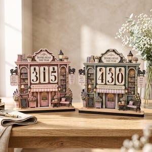 May include: Two decorative bookstore-themed book trackers with rotating number displays. The left unit is pink, the right is green. Both feature the text 'BOOKS READ THIS YEAR', 'SOPHIA'S BOOKSTORE', and small signs reading 'Every book is a new adventure'.