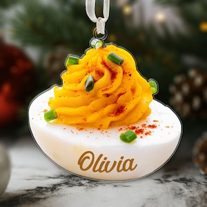 Peut inclure: Ornement de Noël en forme d'œuf mimosa, avec une base d'œuf blanche et un tourbillon de garniture jaune, garni d'oignons verts et de paprika. Le nom "Olivia" est écrit en cursive sur l'œuf. Un ruban blanc est attaché pour l'accrocher.