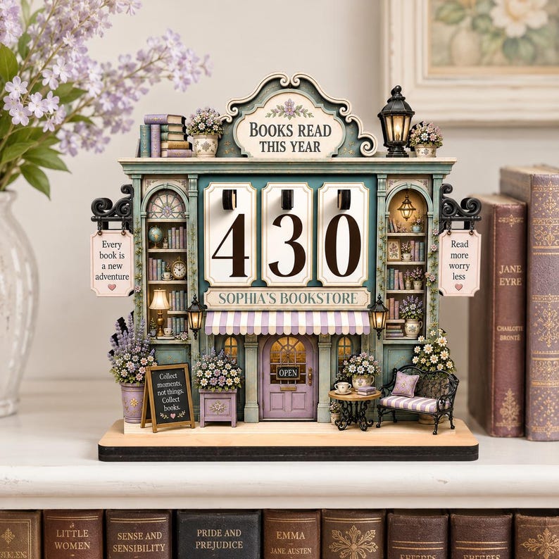 May include: A decorative wooden book tracker shaped like a bookstore with the text BOOKS READ THIS YEAR, SOPHIA'S BOOKSTORE, and numbers 430 displayed. It features miniature shelves, a purple door, a small bench, and floral accents in shades of teal and purple.