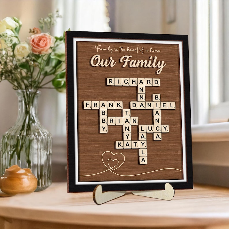 May include: A framed wood sign with a brown background and a white border. The sign features a scrabble tile crossword puzzle with the names "Richard", "Frank", "Daniel", "Brian", "Lucy", and "Katy" spelled out. The sign also includes the text "Family is the heart of a home" and "Our Family".