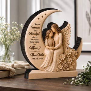 May include: A wooden crescent moon sculpture featuring a carved angel and a child sitting together. The moon is engraved with the text: I know Heaven is a Beautiful place, because they have my Mom, Rose Wild, 04.25.2026. Includes floral accents.