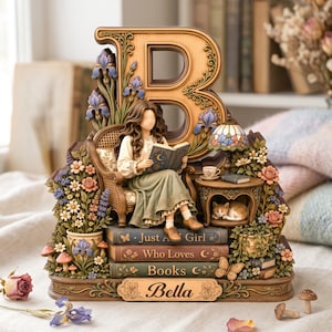Puede incluir: Una figura decorativa con una letra B grande, una persona leyendo un libro y un gato durmiendo. La base incluye libros apilados con las etiquetas Just A Girl, Who Loves, Books y una placa con el nombre Bella, rodeada de motivos florales.