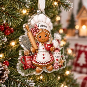 May include: A clear acrylic Christmas ornament shaped like a gingerbread person wearing a chef hat with the name Emma, a red and white checkered apron, and holding a rolling pin and red mug. It features a whisk, flour jar, and text Baked With Love.