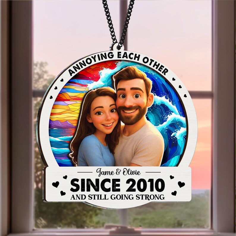 May include: A round, wooden ornament with a cartoon image of a couple, with the text "ANNOYING EACH OTHER" around the top. The bottom has the text "SINCE 2010 AND STILL GOING STRONG". The ornament hangs from a black chain.