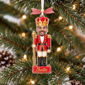 Peut inclure: Une décoration de sapin de Noël en acrylique plat en forme de soldat casse-noisette avec une photo de visage personnalisée, portant un uniforme rouge et or, tenant un bâton doré, avec un nœud vichy rouge et blanc et le nom Justin inscrit à la base.