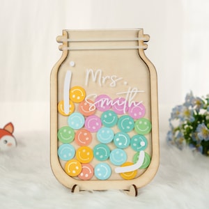 May include: A wooden jar-shaped sign with the name "Mrs. Smith" in white script. The jar is filled with colorful smiley face tokens. The sign is supported by a wooden stand and is a decorative item.