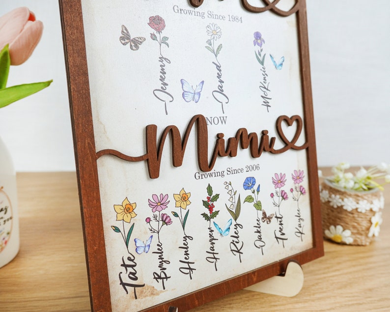 May include: A wooden framed sign with a heart-shaped cutout and the word "Mimi" in brown lettering. The sign features a floral design with names and dates, including "Growing Since 1984" and "Growing Since 2006".
