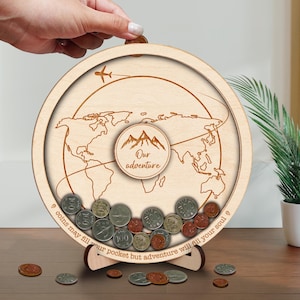 May include: A round wooden travel-themed coin bank with a world map design. The bank features the text "Our adventure" and "coins may fill your pocket but adventure will fill your soul". The bank is filled with various coins and is displayed on a wooden surface.