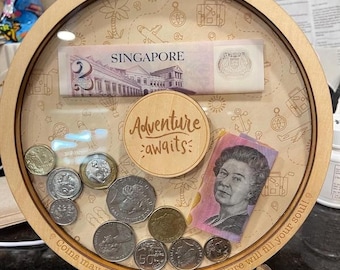 May include: A wooden coin bank with a glass front, featuring a Singapore dollar bill, coins, and a sign that reads "Adventure awaits". The bank has a circular base and a quote around the edge that reads "Coins may fill your pocket but adventure will fill your soul!"