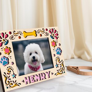 May include: A wooden photo frame with a picture of a white dog wearing a pink bandana. The frame is decorated with colorful floral and paw print cutouts, and the name "HENRY" is displayed at the bottom. A dog collar rests beside the frame.