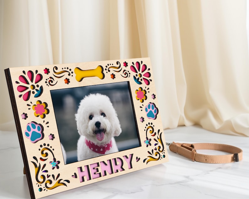May include: A wooden photo frame with a picture of a white dog wearing a pink bandana. The frame is decorated with colorful floral and paw print cutouts, and the name "HENRY" is displayed at the bottom. A dog collar rests beside the frame.