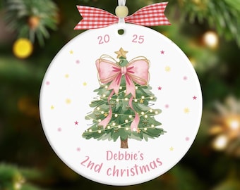 May include: A white ceramic Christmas ornament with a hand-drawn Christmas tree design. The tree is decorated with a pink bow, lights, and a star. The ornament has the text "Debbie's 2nd Christmas" and the year "2025". A red and white checkered bow is at the top.