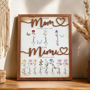 May include: A brown wooden framed sign with the words "FIRST Mom Growing Since 1984" and "NOW Mimi Growing Since 2006" in a decorative font. The sign features a heart design and a floral motif with the names of children listed below each flower.
