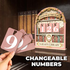 May include: A pink wooden bookshelf-shaped counter with the text "BOOKS READ THIS YEAR" and changeable numbers. The counter displays "126" and includes additional number cards. The text "SARAH'S LIBRARY" is also visible.