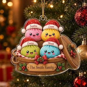 May include: A clear acrylic Christmas ornament featuring four colorful, glittery dumpling characters wearing red Santa hats with the names Sophia, Liam, Emma, and Noah. They sit in a wooden steamer basket labeled The Smith Family with holly.