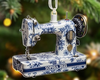 May include: A decorative sewing machine ornament with a white and blue floral pattern. The ornament has a ribbon for hanging and features the text "SOPHIA 2025" on the base. The sewing machine has a gold-colored wheel and accents.