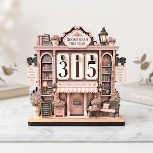 May include: A decorative wooden book tracker shaped like a bookstore facade in shades of pink and brown. It features the text "BOOKS READ THIS YEAR" and "SOPHIA'S BOOKSTORE" with rotating number tiles currently displaying 315, plus miniature floral accents.