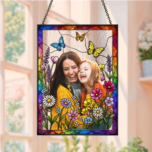 Könnte beinhalten: Buntglasfenster-Kunst mit einem Foto einer lachenden Frau und eines Kindes, umgeben von bunten Blumen und Schmetterlingen. Der Rahmen hat einen Regenbogenrand und wird von einer schwarzen Kette gehalten.