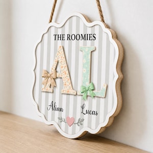 Könnte beinhalten: Ein gewelltes Holzschild für die Wand mit grau-weiß gestreiftem Hintergrund. Es zeigt den Text THE ROOMIES, die Buchstaben A und L mit Blumenmustern und Schleifen, die Namen Alian und Lucas sowie ein kleines rosa Herz mit grauen Blättern.