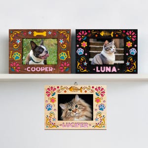 May include: Three decorative pet photo frames. The frames feature colorful floral and paw print designs. One frame has a dog named "COOPER", and the other two have cats named "LUNA" and "LUCIFER". The frames are made of wood.