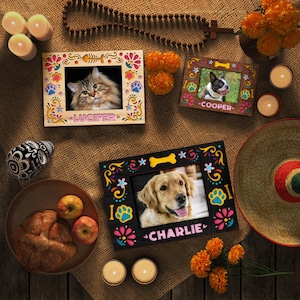 May include: A collection of decorative wooden picture frames with colorful floral and paw print designs. The frames hold photos of a cat and two dogs, with names like "Lucifer" and "Charlie" displayed. The scene includes candles, flowers, and a sombrero.