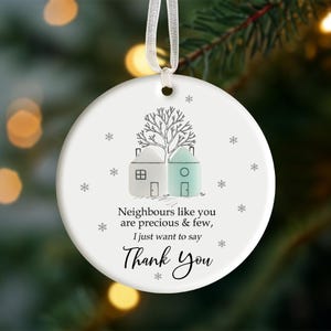 Könnte beinhalten: Runder weißer Keramik-Weihnachtsschmuck mit einer Zeichnung von zwei Häusern und einem Baum. Der Text lautet: „Nachbarn wie Sie sind kostbar und rar, ich möchte einfach nur Danke sagen“. Hängt an einem weißen Band.