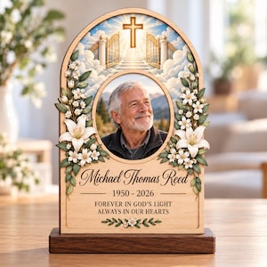 May include: An arched wooden memorial plaque featuring a circular photo of an older man, flanked by white lilies and green leaves. The top shows a cross and golden gates in the clouds. Text reads: Michael Thomas Reed, 1950-2026, FOREVER IN GOD'S LIGHT ALWAYS IN OUR HEARTS.