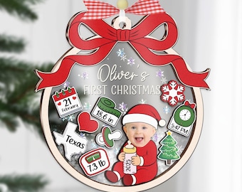 Op de afbeelding: Een gepersonaliseerde kerstbal met een babyfoto in een rood kerstpak, met de tekst "Oliver's First Christmas". Inclusief geboortegegevens: 47.2 cm, 3.3 kg, en de datum. De ornament heeft een rode strik en een wit lint om op te hangen.