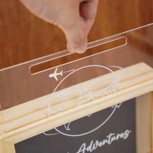 May include: A wooden shadow box with a clear acrylic lid. The lid has a slot for inserting money and features a world map and airplane design. The box has a black chalkboard insert with the text "Our Adventures" written on it.