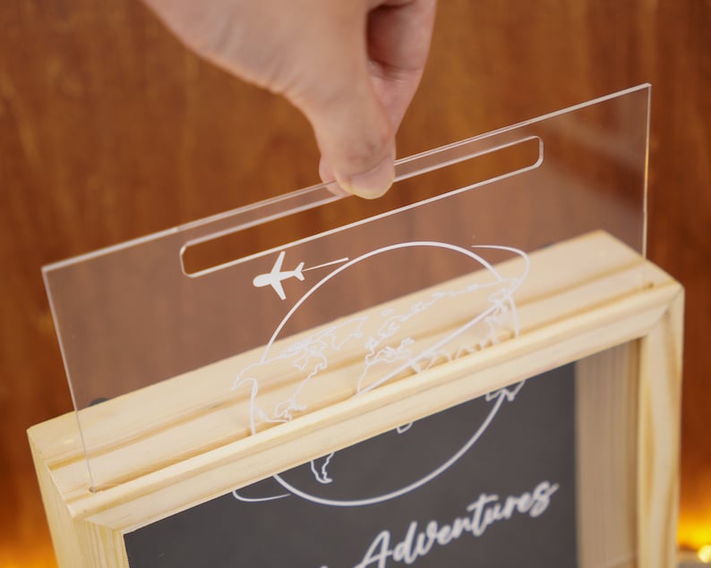 May include: A wooden shadow box with a clear acrylic lid. The lid has a slot for inserting money and features a world map and airplane design. The box has a black chalkboard insert with the text "Our Adventures" written on it.