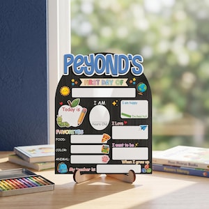 May include: A black chalkboard-style sign with the text "PEYOND'S FIRST DAY OF" in blue and white. The sign includes sections for "Today is," "Favorites," and "I am." It has a wooden stand and is designed for a child's first day of school.