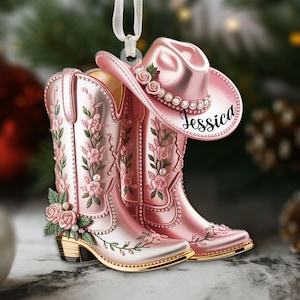 Puede incluir: Un adorno navideño rosa con un par de botas de vaquero y un sombrero de vaquero. Las botas y el sombrero están decorados con diseños florales y perlas. El sombrero tiene el nombre "Jessica".