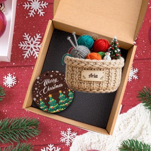 Puede incluir: Una caja de regalo con tem&aacute;tica navide&ntilde;a con un adorno de madera y una cesta de hilo. El adorno dice "Merry Christmas" y presenta &aacute;rboles de Navidad verdes. La cesta de hilo contiene bolas de hilo de colores, agujas de tejer y un peque&ntilde;o &aacute;rbol de Navidad. La cesta tiene el nombre "Aria".