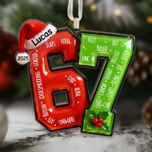 May include: A festive Christmas ornament in the shape of the numbers "67", with a red "6" and a green "7". The "6" features a Santa hat with the name "Lucas" and the year "2025". Slang terms are written on the ornament.