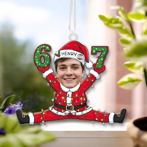 May include: A personalized Christmas ornament featuring a person's face in a Santa Claus suit, holding the numbers 6 and 7. The ornament is red with a gold belt, and the numbers are green with white polka dots. The name "Henry" is on the hat.