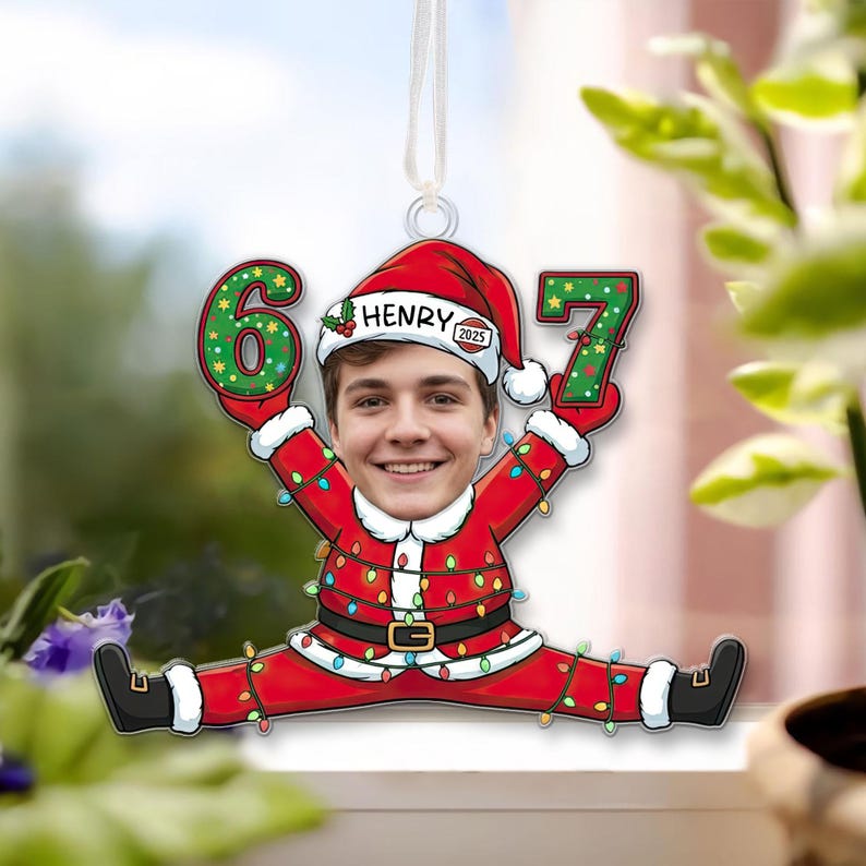 May include: A personalized Christmas ornament featuring a person's face in a Santa Claus suit, holding the numbers 6 and 7. The ornament is red with a gold belt, and the numbers are green with white polka dots. The name "Henry" is on the hat.