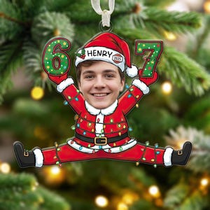 May include: A personalized Christmas ornament featuring a person's face in a Santa Claus outfit, holding the numbers 6 and 7. The ornament has a red and white color scheme with a string of colorful lights and the name "Henry" on the hat.