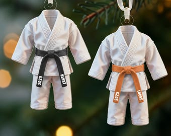 May include: Two white karate uniform ornaments with black and orange belts. The black belt has "2025" and "HENRY" printed on it. The orange belt has "2025" and "LUCAS" printed on it. Each ornament has a white ribbon for hanging.