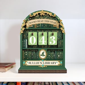 May include: A green wooden bookshelf-shaped counter with the text "BOOKS READ THIS YEAR" and "043" displayed on green cards. The bottom of the counter reads "JULLIE'S LIBRARY". The bookshelf is decorated with gold accents and a witch hat.