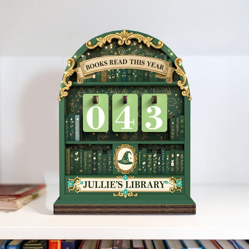 May include: A green wooden bookshelf-shaped counter with the text "BOOKS READ THIS YEAR" and "043" displayed on green cards. The bottom of the counter reads "JULLIE'S LIBRARY". The bookshelf is decorated with gold accents and a witch hat.