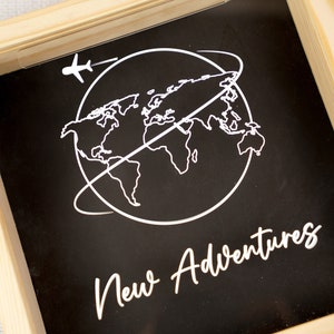 May include: A wooden frame with a black background and a white outline of the world with an airplane flying above it. The text "New Adventures" is written in white cursive below the world.