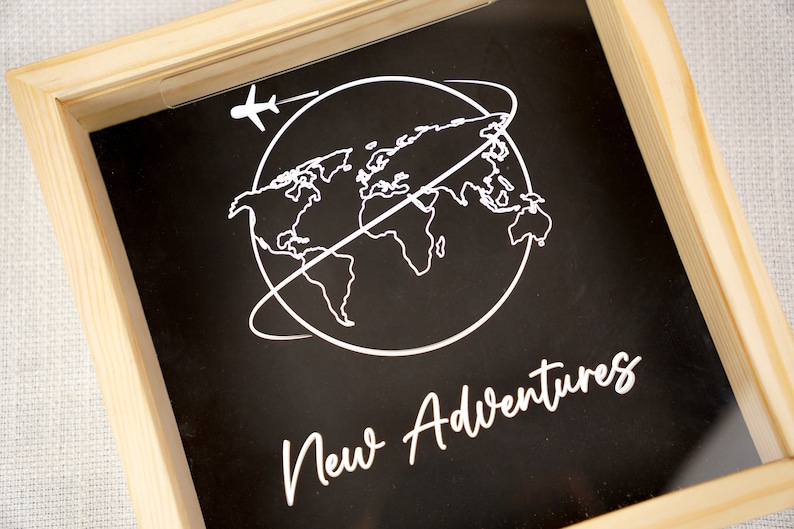 May include: A wooden frame with a black background and a white outline of the world with an airplane flying above it. The text "New Adventures" is written in white cursive below the world.