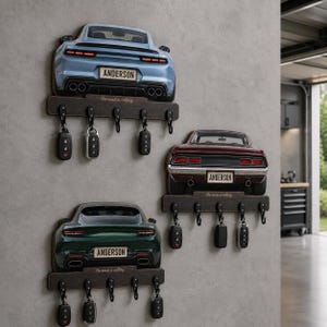 Könnte beinhalten: Drei Wand-Schlüsselhalter mit Heckansichten von Sportwagen in Hellblau, Dunkelgrün und Kastanienbraun. Jedes Holzregal hat fünf schwarze Haken und ein Nummernschild mit der Aufschrift ANDERSON. Der Text The road is calling ist auf jedem Regal gedruckt.