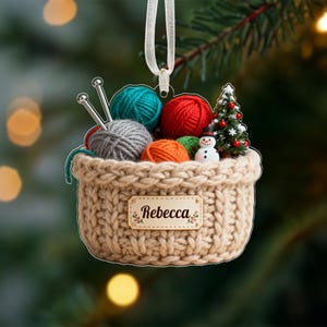 Puede incluir: Adorno navide&ntilde;o en forma de cesta tejida de color beige, llena de bolas de hilo de colores, agujas de tejer, un &aacute;rbol de Navidad en miniatura y un mu&ntilde;eco de nieve. El adorno tiene una cinta blanca para colgar y una etiqueta que dice "Rebecca".